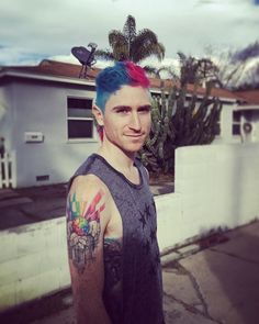 a man with colorful hair and tattoos standing in front of a white fence next to a house