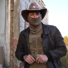 a man wearing a mask and jacket standing in front of a building with a brick wall