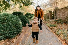 a little boy is walking down the sidewalk with his mother in the background and leaves on the ground