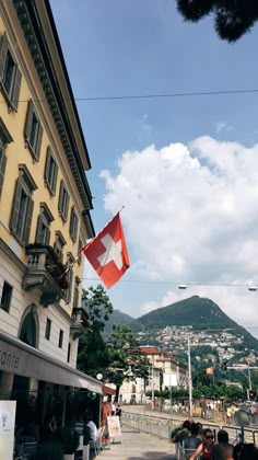 a flag flying in the air next to a building with people sitting on benches near it