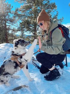 a woman kneeling down next to a dog on top of snow covered ground with trees in the background