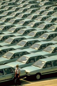 rows of parked cars in a parking lot with one man walking past them and the other person looking at it