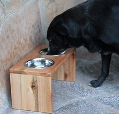 a black dog eating food out of a wooden elevated bowl on top of a table