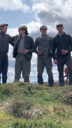 four men standing on top of a grass covered hill next to each other wearing hats and ties