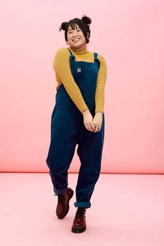 a woman standing in front of a pink background wearing blue overalls and a yellow sweater