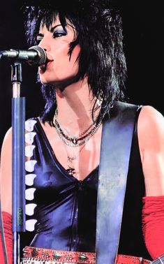 a woman with black hair holding a guitar in front of a microphone and wearing red gloves