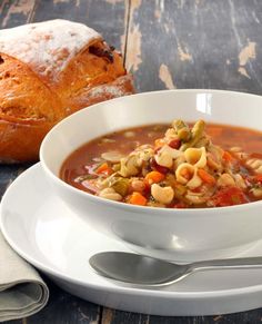 a white bowl filled with pasta soup next to a loaf of bread on a table