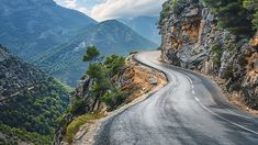 an empty road winding through the mountains with trees on both sides and clouds in the sky