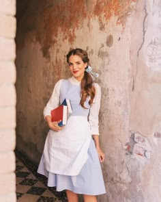 a woman in an apron and dress is standing by a wall with books on her shoulder