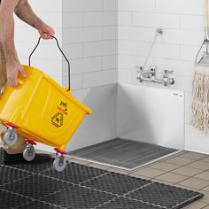 a man pushing a yellow bucket on wheels in a bathroom with white tile walls and flooring