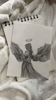 two drawings of an angel on top of a white blanket next to a spiral notebook