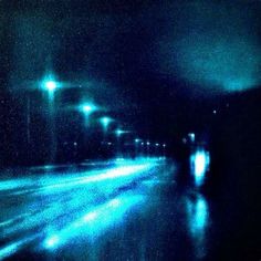 blurry photograph of street lights at night with rain on the ground and one car driving down the road