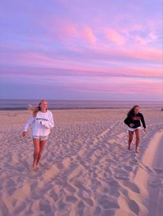 two girls are running in the sand on the beach at sunset, one is wearing a white shirt