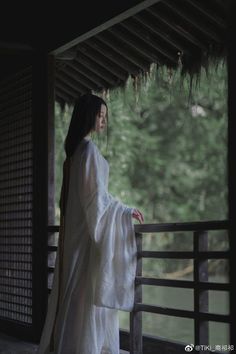 a woman dressed in white standing on a porch next to a tree and water area