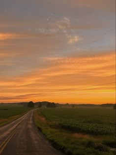 an empty road in the middle of a green field at sunset with clouds above it