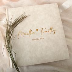 a wedding guest book with gold lettering on it and a plant in the corner next to it