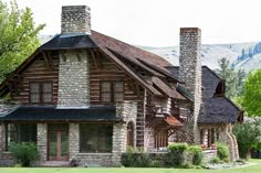 an old log house with stone chimneys in the front yard and mountains in the background