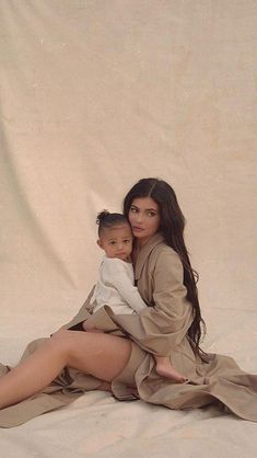 a woman holding a baby sitting on top of a white sheet covered floor next to a wall