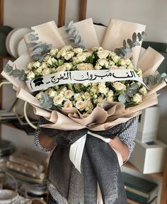 a bouquet of white roses with an arabic writing on the ribbon is held by someone's hand