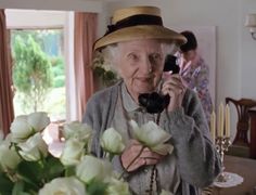 an old woman is talking on the phone with flowers in front of her and people standing around