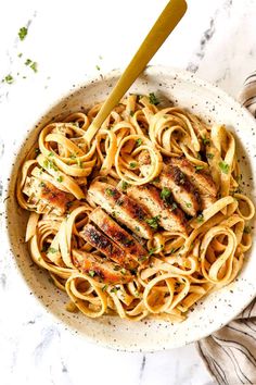 a white bowl filled with chicken and pasta on top of a marble countertop next to a wooden spoon