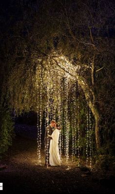 a bride and groom standing under an archway covered with lights at night in the woods