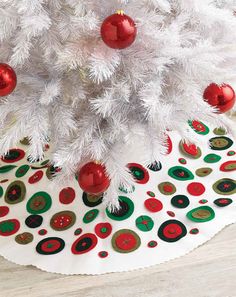 a white christmas tree with red and green ornaments on it's base, sitting in front of a wooden table