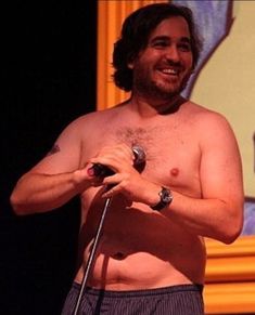 a shirtless man holding a microphone in his right hand and smiling at the camera