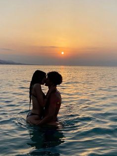 two people are sitting in the water at sunset, one is kissing the other's forehead