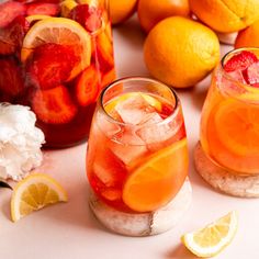 two glasses filled with fruit and ice next to sliced lemons, strawberries, and watermelon