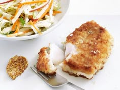 a white plate topped with fish next to a bowl of coleslaw and a fork