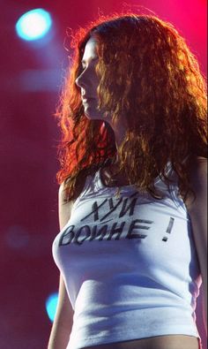 a woman with long red hair standing in front of a microphone and wearing a white tank top