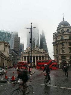 people riding bikes in the rain on a city street with double decker buses and tall buildings