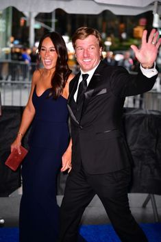 a man in a tuxedo waves to the camera while standing next to a woman on a blue carpet