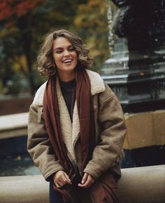 a woman is sitting on a bench in front of a fountain and smiling at the camera