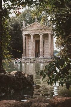 an old building sitting in the middle of a lake surrounded by rocks and trees with people standing around it