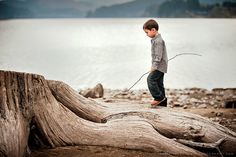 a young boy standing on top of a tree stump next to the ocean with a stick in his hand