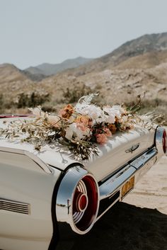 the back end of an old car with flowers on it's hood, in front of mountains