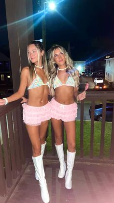 two beautiful young women standing next to each other on a balcony at night with lights in the background