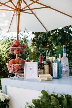 an outdoor bar set up with drinks and fruit on the top shelf, under an umbrella