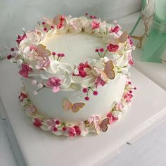 a white cake with pink flowers and butterflies on it sitting on a table next to a green box