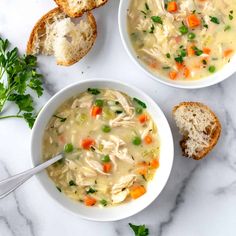 two bowls of chicken noodle soup on a marble table with bread and parsley