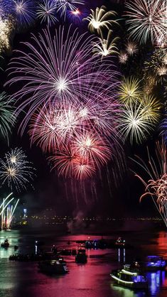 fireworks are lit up in the night sky over water and boats on the river below