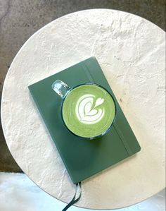 a cup of green coffee sitting on top of a white table next to a book