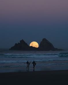 two people are walking on the beach with their surfboards in front of the moon