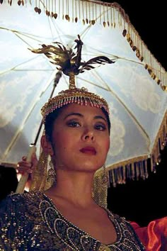 a woman in an elaborate headdress holds an umbrella over her head and looks off to the side