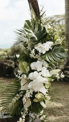 white flowers and greenery are arranged on a wooden stand in the grass near palm trees