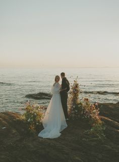 a bride and groom standing on the rocks by the water at sunset in their wedding attire