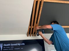 a man in blue shirt working on a ceiling with wooden rafters above him and tv screen behind him