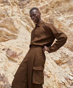 a man standing in front of a mountain wearing a brown shirt and wide legged pants